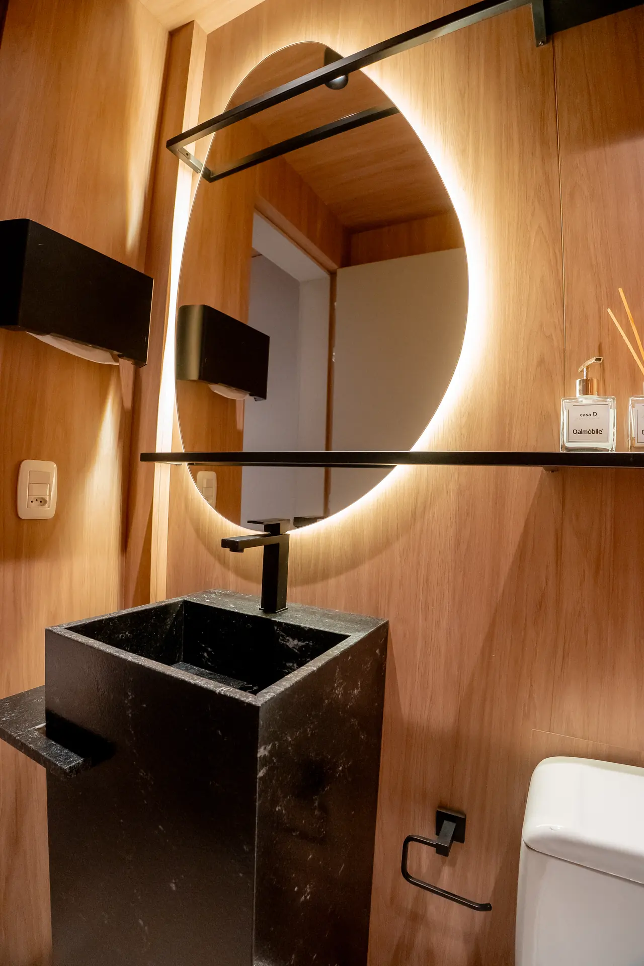 Detalhe de lavabo com espelho circular retroiluminado e marcenaria amadeirada de piso a teto.