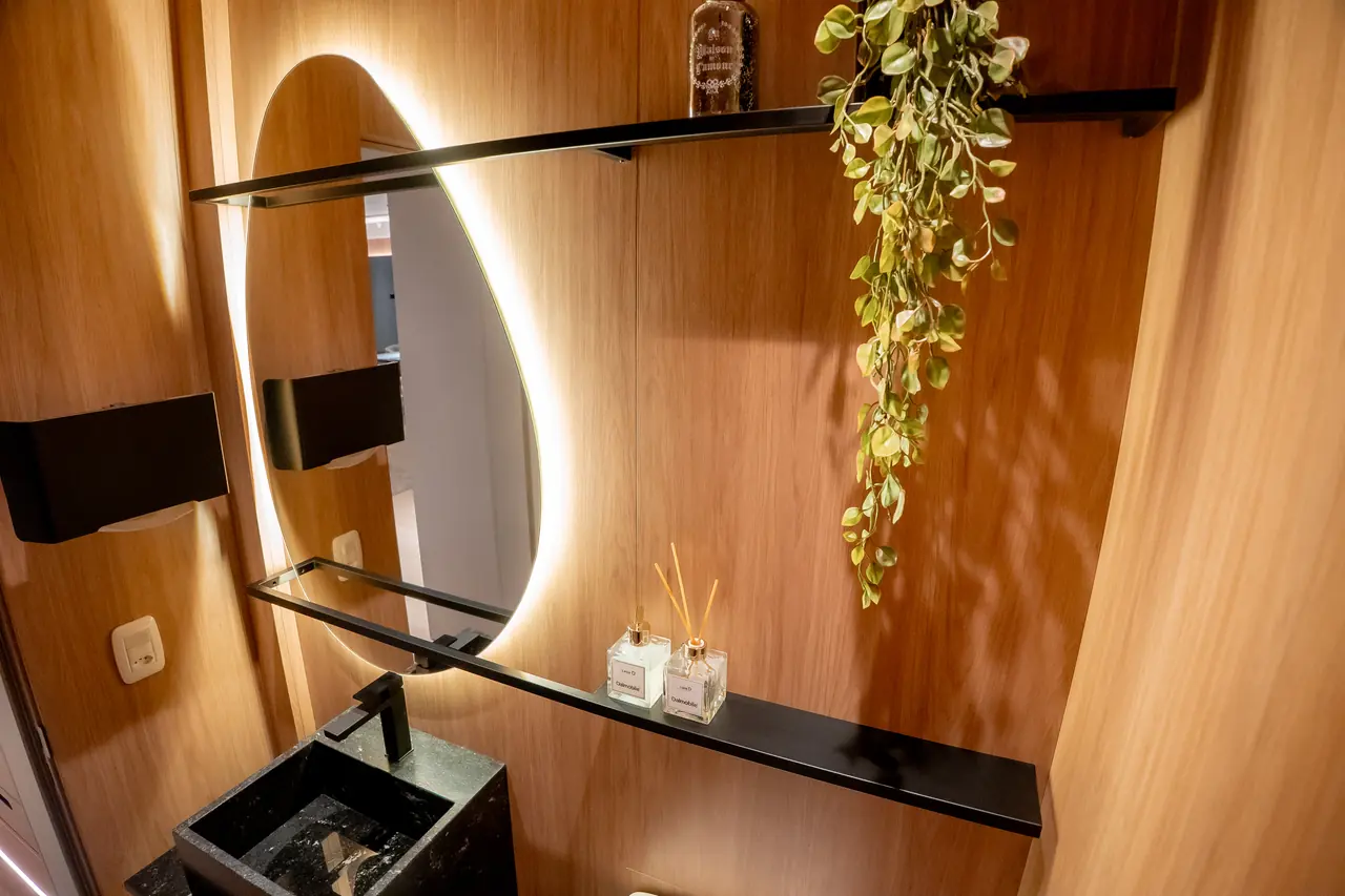 Lavabo revestido em madeira com espelho orgânico iluminado, cuba esculpida e prateleira metálica.
