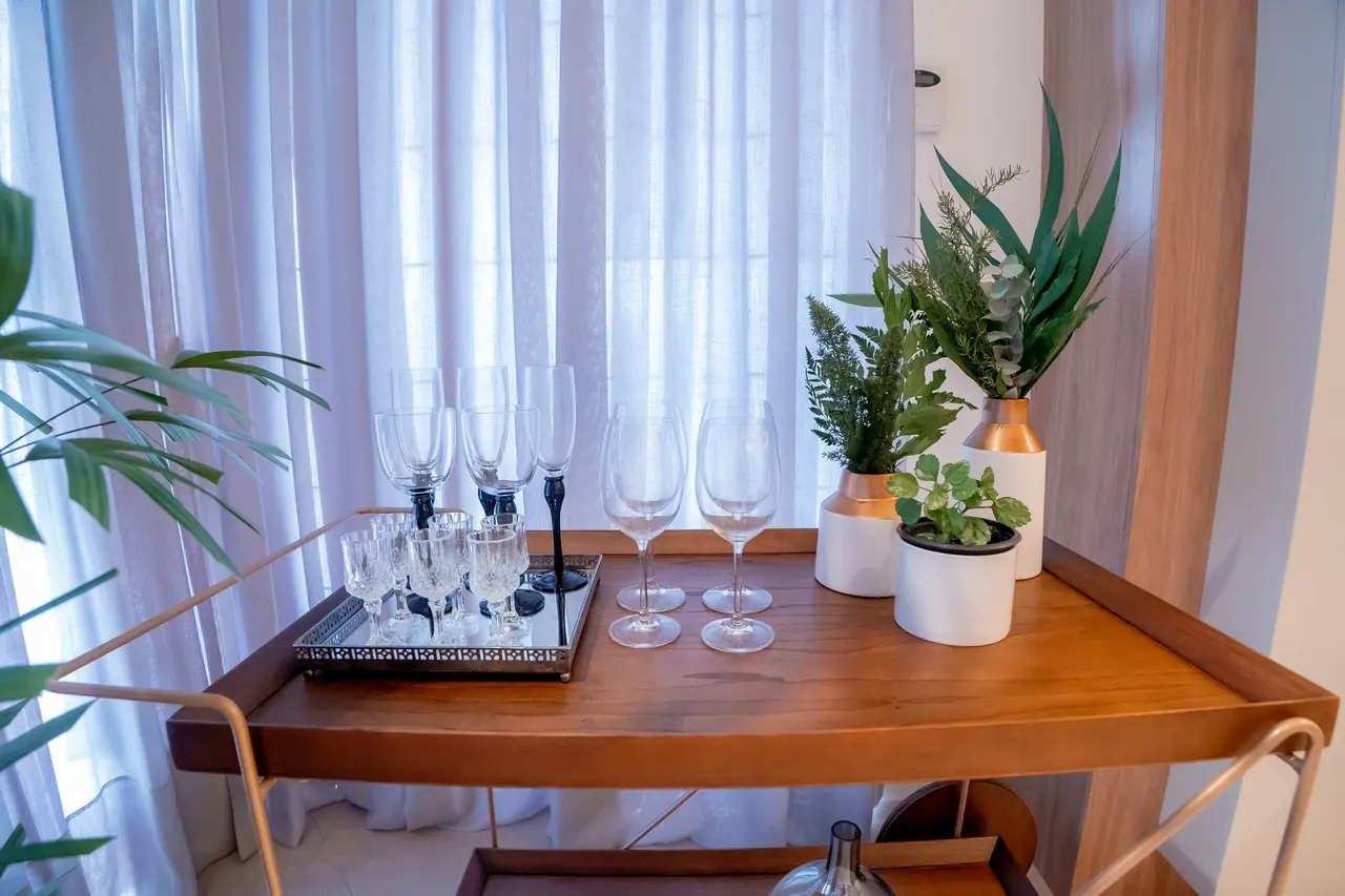 Cantinho gourmet com carrinho bar em madeira, taças e plantas ao lado de cortinas de linho.
